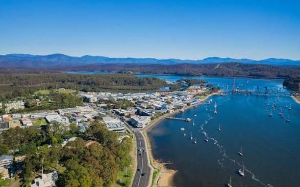 Aerial view of Batemans Bay on a clear day: the town along the Clyde River, yachts moored on the water, the bridge upstream and the mountains behind